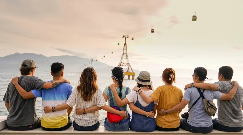 Seven friends sitting in a row with their arms around each other, looking out over the water