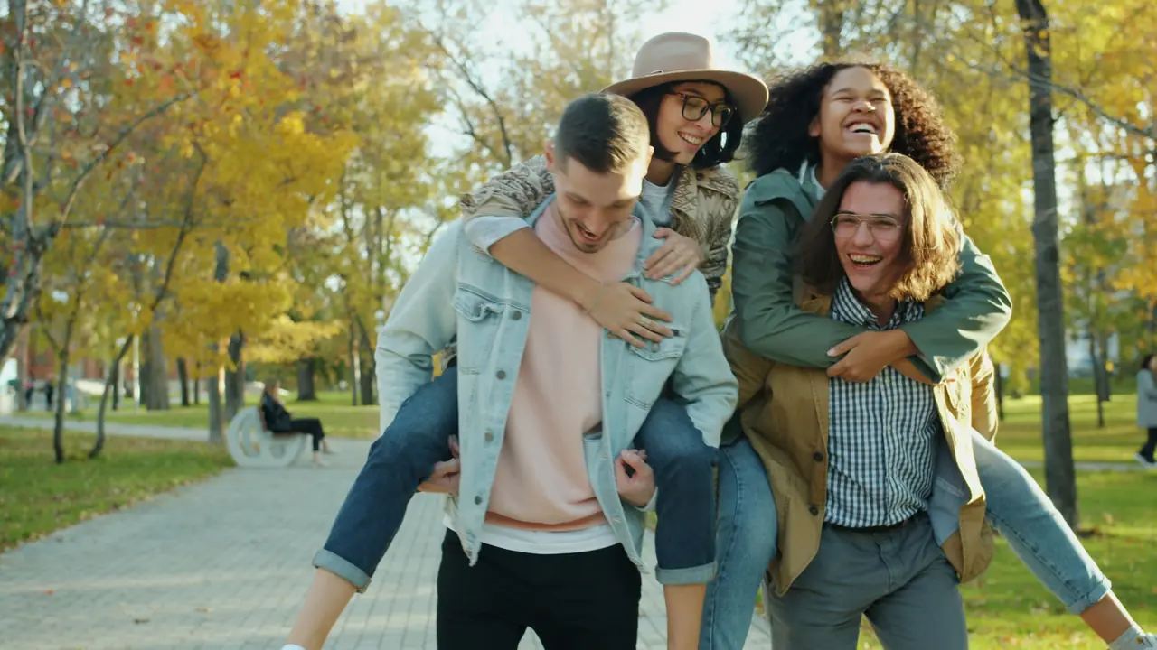 Friends laughing as two of them get piggyback rides in an autumn park