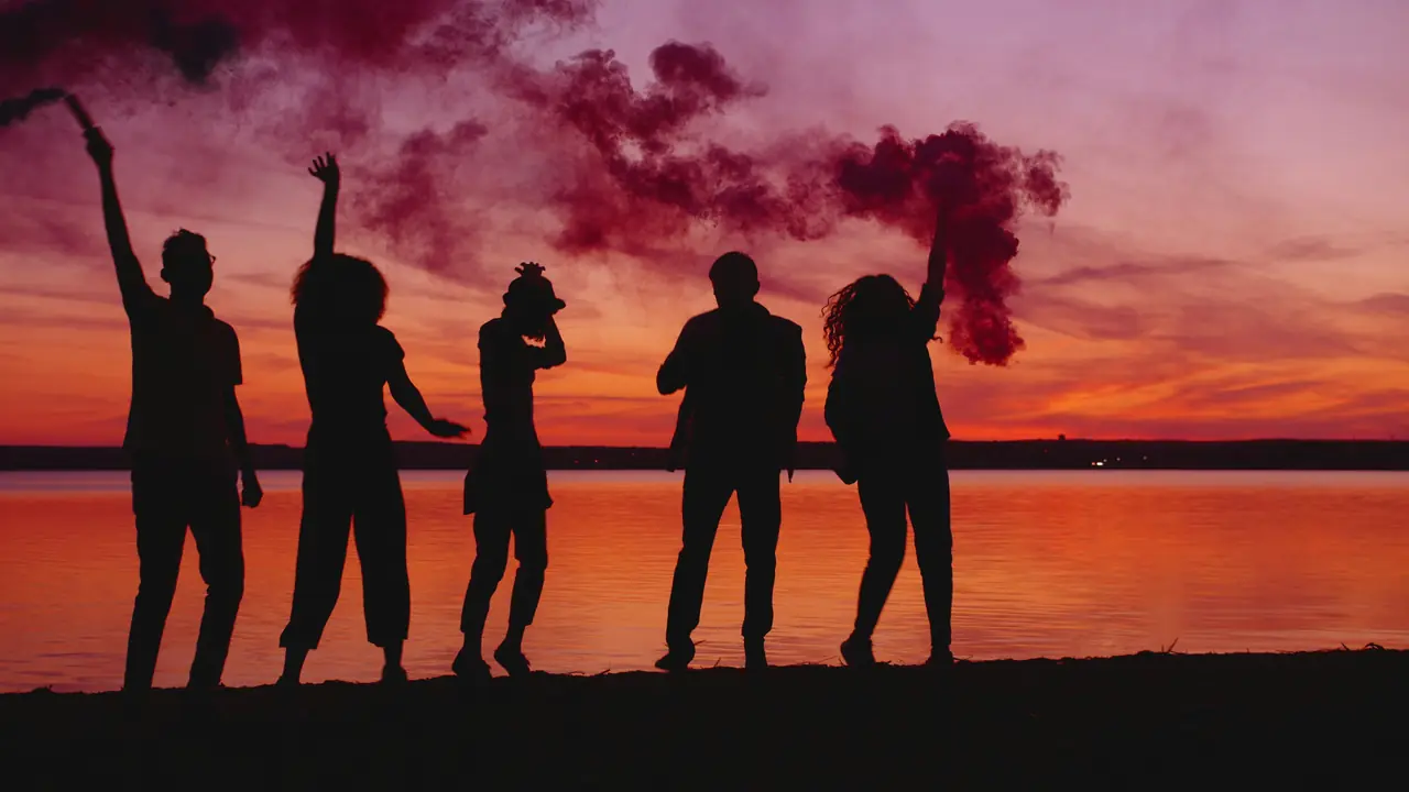 Five friends silhouetted against a pink and orange sunset, one holding up red smoke